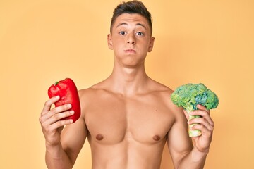 Young hispanic boy shirtless holding broccoli and red pepper puffing cheeks with funny face. mouth inflated with air, catching air.