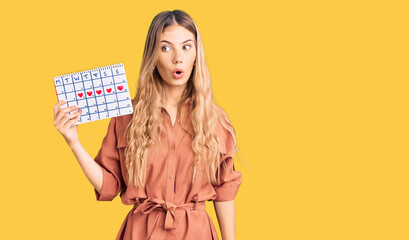 Beautiful caucasian woman with blonde hair holding heart calendar scared and amazed with open mouth for surprise, disbelief face