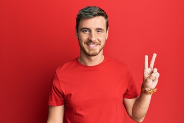 Naklejka premium Handsome caucasian man wearing casual red tshirt smiling looking to the camera showing fingers doing victory sign. number two.