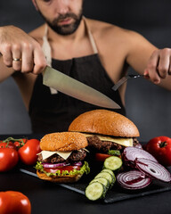 A man with Cutlery eats a huge hamburger on a dark background.