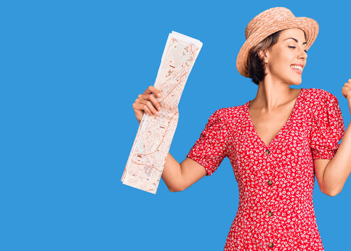 Young beautiful woman wearing summer hat holding map pointing thumb up to the side smiling happy with open mouth