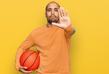 Hispanic adult man holding basketball ball with open hand doing stop sign with serious and confident expression, defense gesture