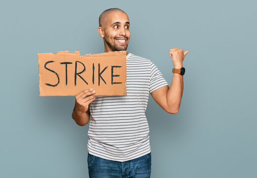 Hispanic adult man holding strike banner cardboard pointing thumb up to the side smiling happy with open mouth