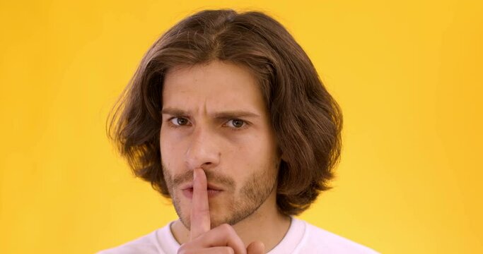 Young mysterious guy putting finger on lips and shaking head, asking for privacy and silence, orange studio background