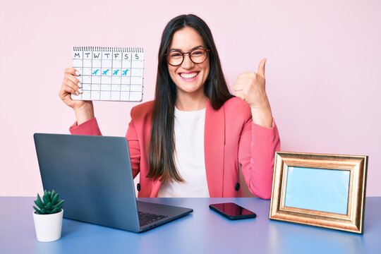Young caucasian woman sitting at the desk holding travel calendar smiling happy and positive, thumb up doing excellent and approval sign