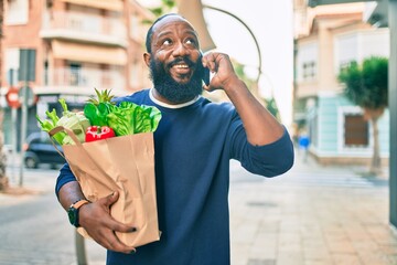 African american man with beard holding paper bag of groceries from supermarket speaking on the...