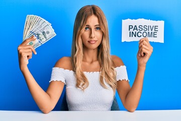 Beautiful blonde young woman holding dollars and passive income text relaxed with serious...