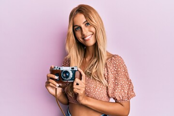 Beautiful blonde young woman holding vintage camera smiling with a happy and cool smile on face....