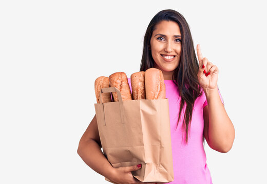 Young beautiful brunette woman holding delivery bag with bread surprised with an idea or question pointing finger with happy face, number one