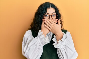 Young brunette woman with curly hair wearing casual clothes and glasses shocked covering mouth with hands for mistake. secret concept.