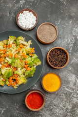 Vertical view of vegetable meal with brocoli and carrots on a black plate and spices on gray table
