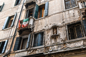 Fototapeta na wymiar The flags of Italy and the city of Venice fly from a balcony window in a rundown, decaying building in the center of Venice, Italy.