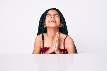 Young african american girl child with braids wearing casual clothes sitting on the table begging and praying with hands together with hope expression on face very emotional and worried. begging.