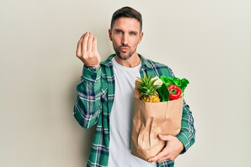 Handsome man with beard holding paper bag with groceries doing italian gesture with hand and...