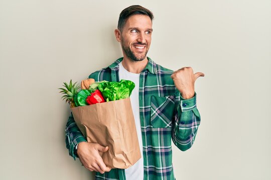 Handsome man with beard holding paper bag with groceries smiling with happy face looking and pointing to the side with thumb up.