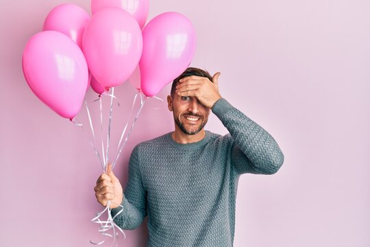 Handsome man with beard holding pink balloons stressed and frustrated with hand on head, surprised and angry face