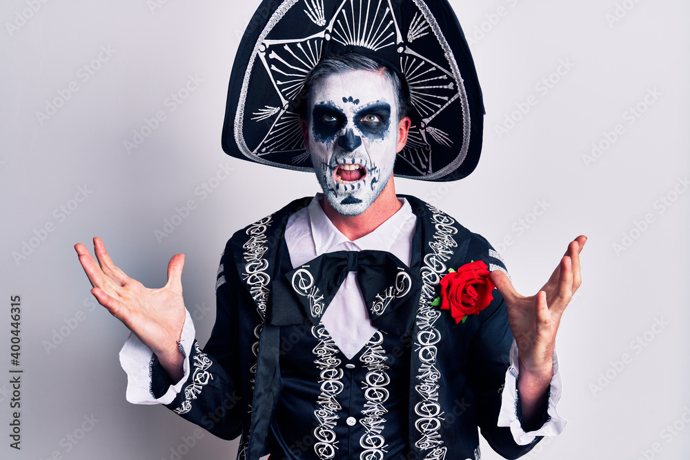 Poster young man wearing mexican day of the dead costume over white crazy and mad shouting and yelling with