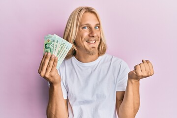 Caucasian young man with long hair holding 20 thai baht banknotes screaming proud, celebrating victory and success very excited with raised arm