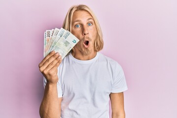 Caucasian young man with long hair holding czech koruna banknotes scared and amazed with open mouth for surprise, disbelief face