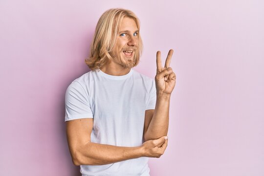 Caucasian young man with long hair wearing casual white t shirt smiling with happy face winking at the camera doing victory sign. number two.