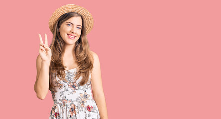 Beautiful caucasian young woman wearing summer hat showing and pointing up with fingers number two while smiling confident and happy.