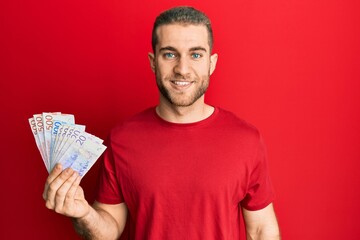 Young caucasian man holding swedish krona banknotes looking positive and happy standing and smiling with a confident smile showing teeth