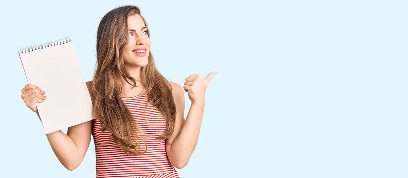 Beautiful caucasian young woman holding notebook pointing thumb up to the side smiling happy with open mouth