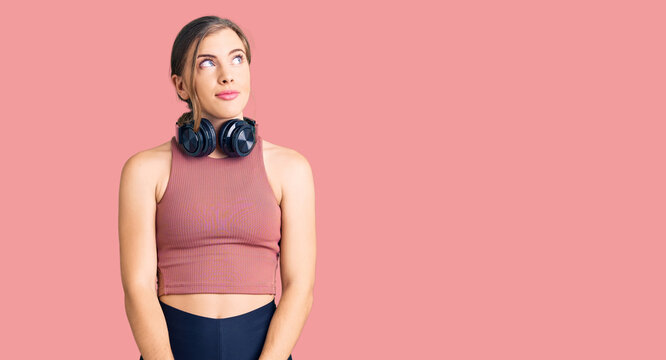 Beautiful caucasian young woman wearing gym clothes and using headphones smiling looking to the side and staring away thinking.
