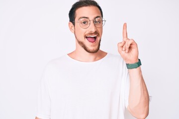Naklejka premium Young handsome man wearing casual clothes and glasses smiling with an idea or question pointing finger up with happy face, number one
