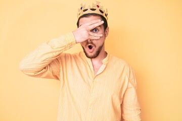 Young handsome man wearing king crown peeking in shock covering face and eyes with hand, looking through fingers afraid