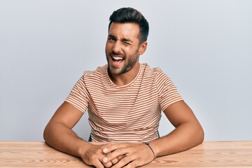 Handsome hispanic man wearing casual clothes sitting on the table winking looking at the camera...