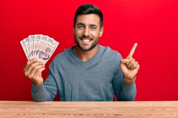Handsome hispanic man holding mexican pesos smiling happy pointing with hand and finger to the side - obrazy, fototapety, plakaty
