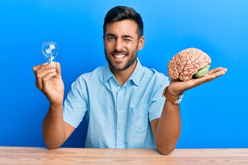 Handsome hispanic man holding lightbulb and brain for inspiration and idea winking looking at the...