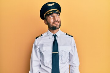 Handsome hispanic man wearing airplane pilot uniform smiling looking to the side and staring away thinking.