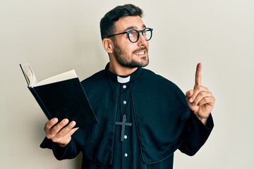 Young hispanic priest man holding bible with finger up smiling looking to the side and staring away...