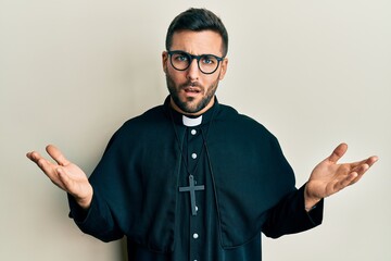 Young hispanic priest man standing over white background in shock face, looking skeptical and...