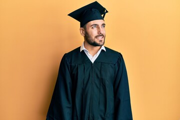 Young hispanic man wearing graduation cap and ceremony robe looking away to side with smile on...