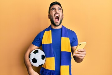 Young hispanic man football hooligan holding soccer ball using smartphone angry and mad screaming...
