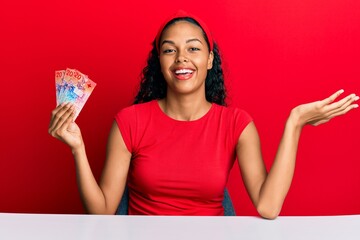 Young african american girl holding swiss franc banknotes sitting on the table celebrating achievement with happy smile and winner expression with raised hand