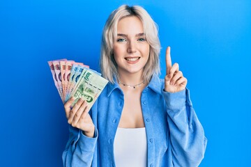 Young blonde girl holding thai baht banknotes smiling with an idea or question pointing finger with happy face, number one