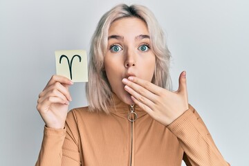 Young blonde girl holding paper with aries zodiac sign covering mouth with hand, shocked and afraid...