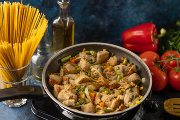 Pan with fragrant frying meat and mix of vegetables on the stove near ingredients on dark blue background. Backstage of cooking pasta. Traditional italian cuisine. Cooking process. Cookbook.