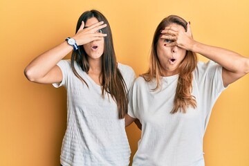 Hispanic family of mother and daughter wearing casual white tshirt peeking in shock covering face and eyes with hand, looking through fingers with embarrassed expression.