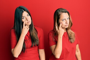 Hispanic family of mother and daughter wearing casual clothes over red background pointing to the eye watching you gesture, suspicious expression