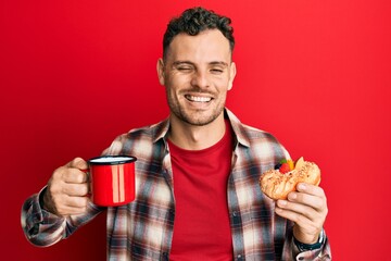 Young hispanic man drinking coffee and eating pastry winking looking at the camera with sexy...