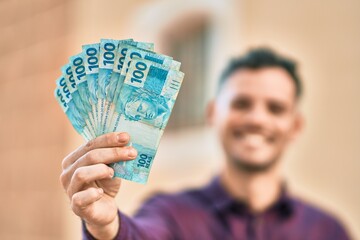 Young hispanic man smiling happy holding brazilian real banknotes at the city.