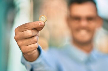 Young hispanic businessman smiling happy holding 1 euro coin at the city.