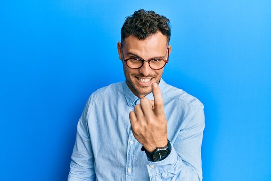 Young hispanic man wearing casual clothes and glasses beckoning come here gesture with hand inviting welcoming happy and smiling