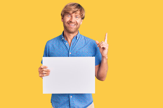 Handsome caucasian man with beard holding sale poster surprised with an idea or question pointing finger with happy face, number one