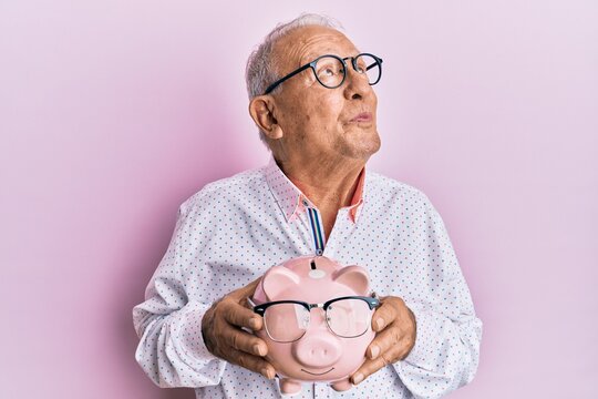 Senior caucasian man holding piggy bank with glasses smiling looking to the side and staring away thinking.